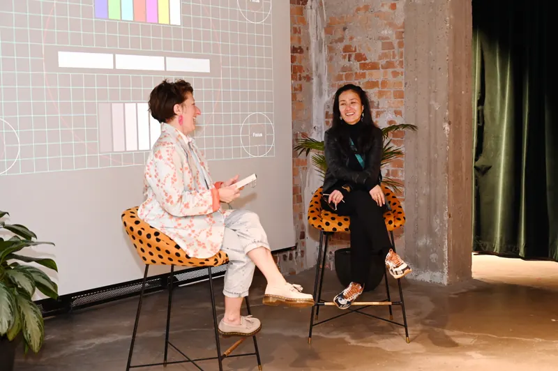 Two speakers seated on stools on stage mid-conversation, a presentation projected behind them
